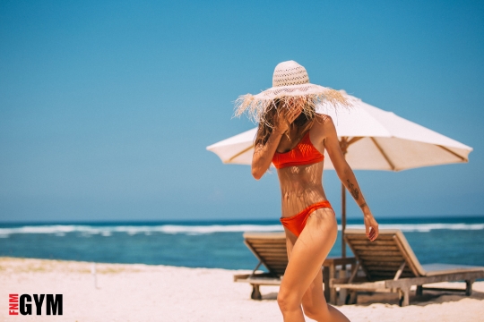 Female in orange bikini walking along the beach with white straw hat on