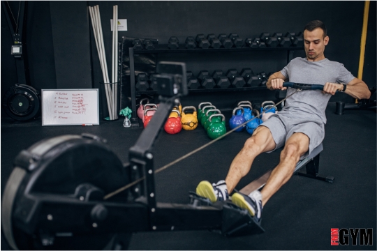 male on rowing machine in gym with kettlebells and dumbbells in view