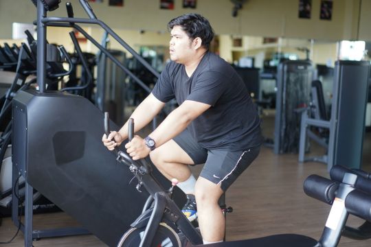 male in black t-shirt riding an exercise bike in gym